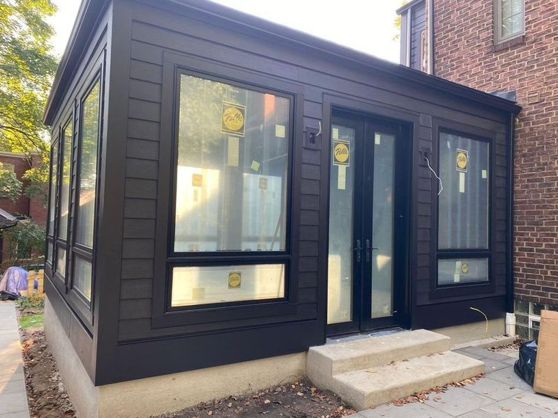 Dark-brown-sided sunroom with large windows, door, and concrete steps. It is attached to a brick building.