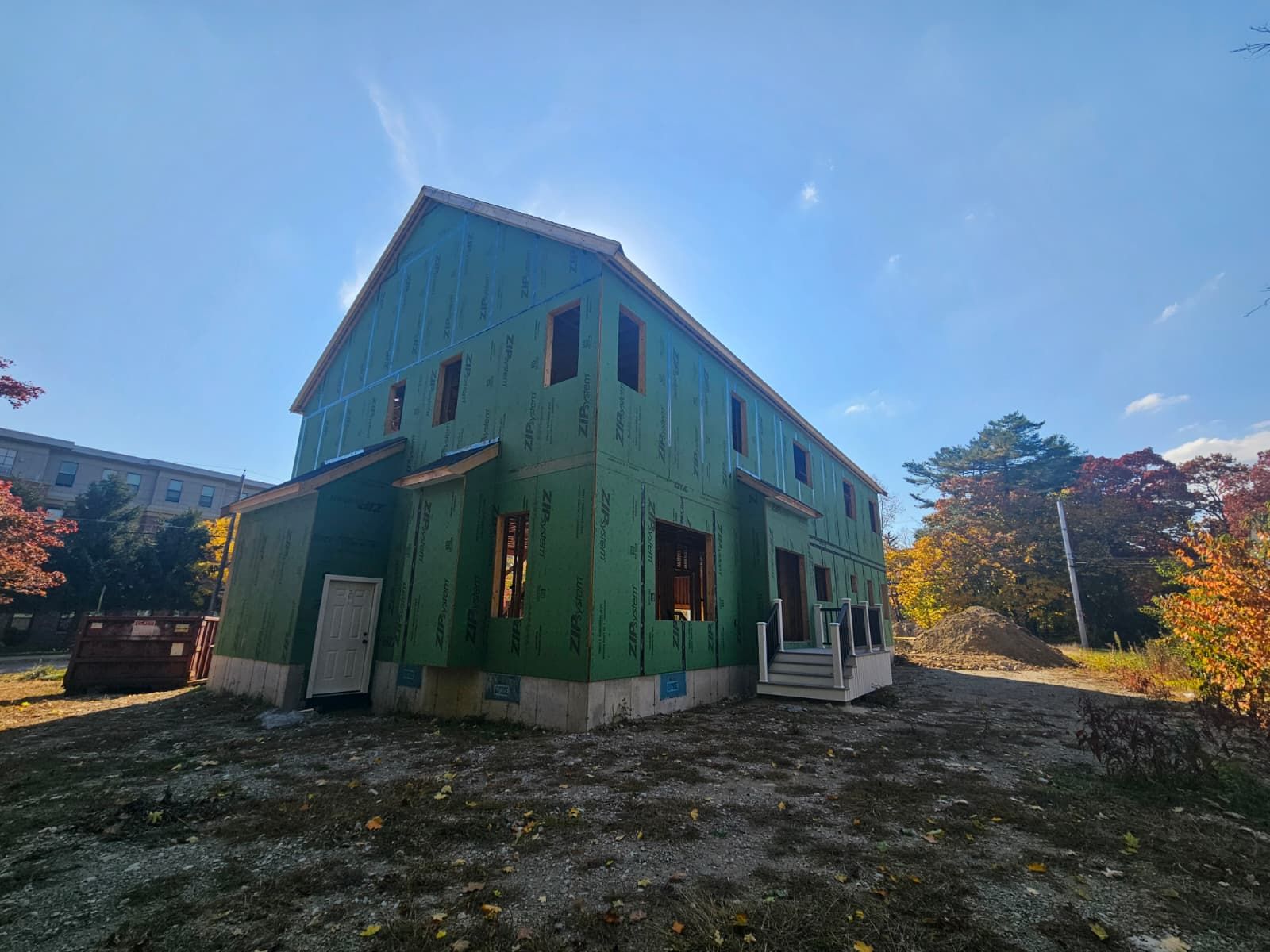 Green barn-style house under a blue sky, surrounded by autumn trees and gravel ground