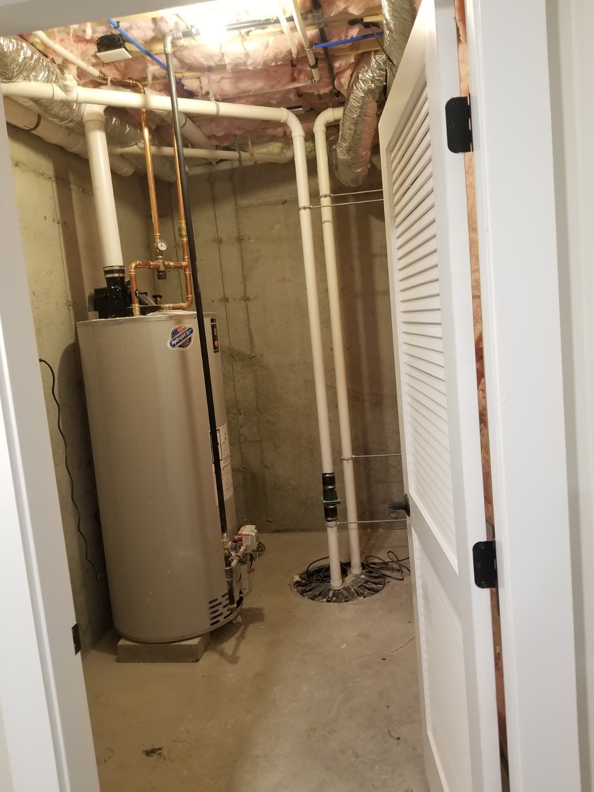 Basement utility room with water heater, exposed pipes, and a floor drain behind a white louvered door