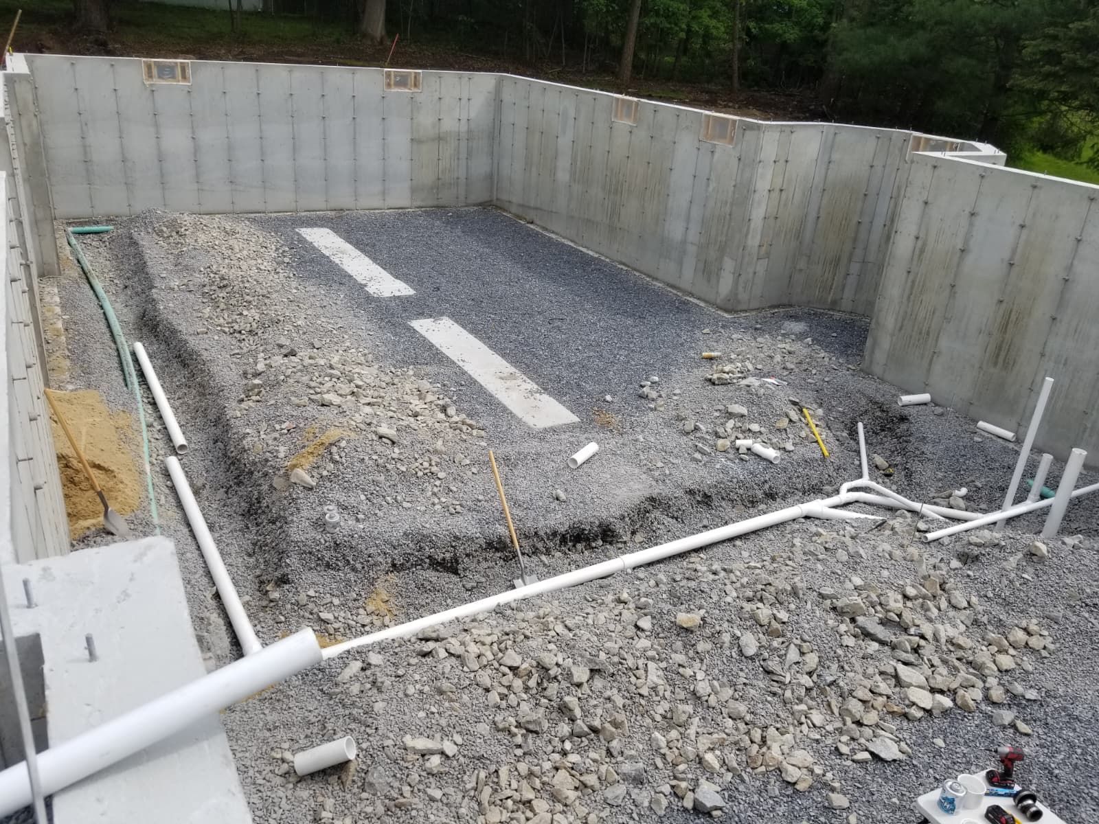 Concrete foundation pit with gravel base and white pipes, surrounded by retaining walls and trees.
