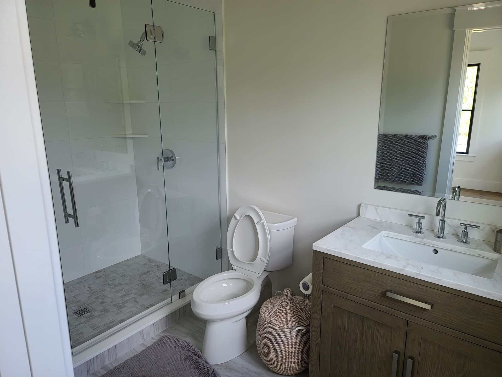 Modern bathroom with glass shower, toilet, vanity sink, mirror, and patterned basket.
