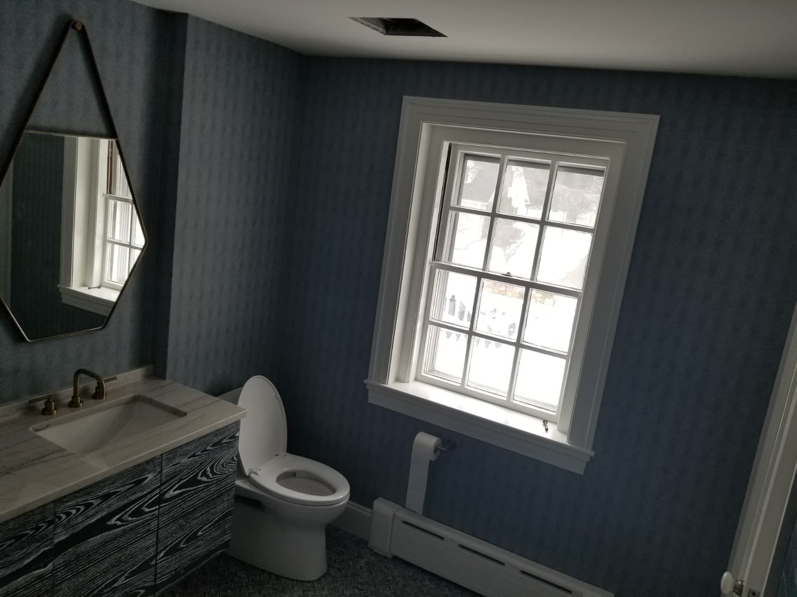 Small bathroom with blue walls, white fixtures, mirror, sink, toilet, and a window with light filtering in