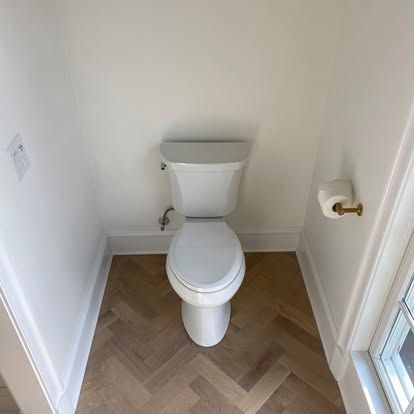 Small bathroom with a white toilet, wood-look floor, and toilet paper holder by a window.