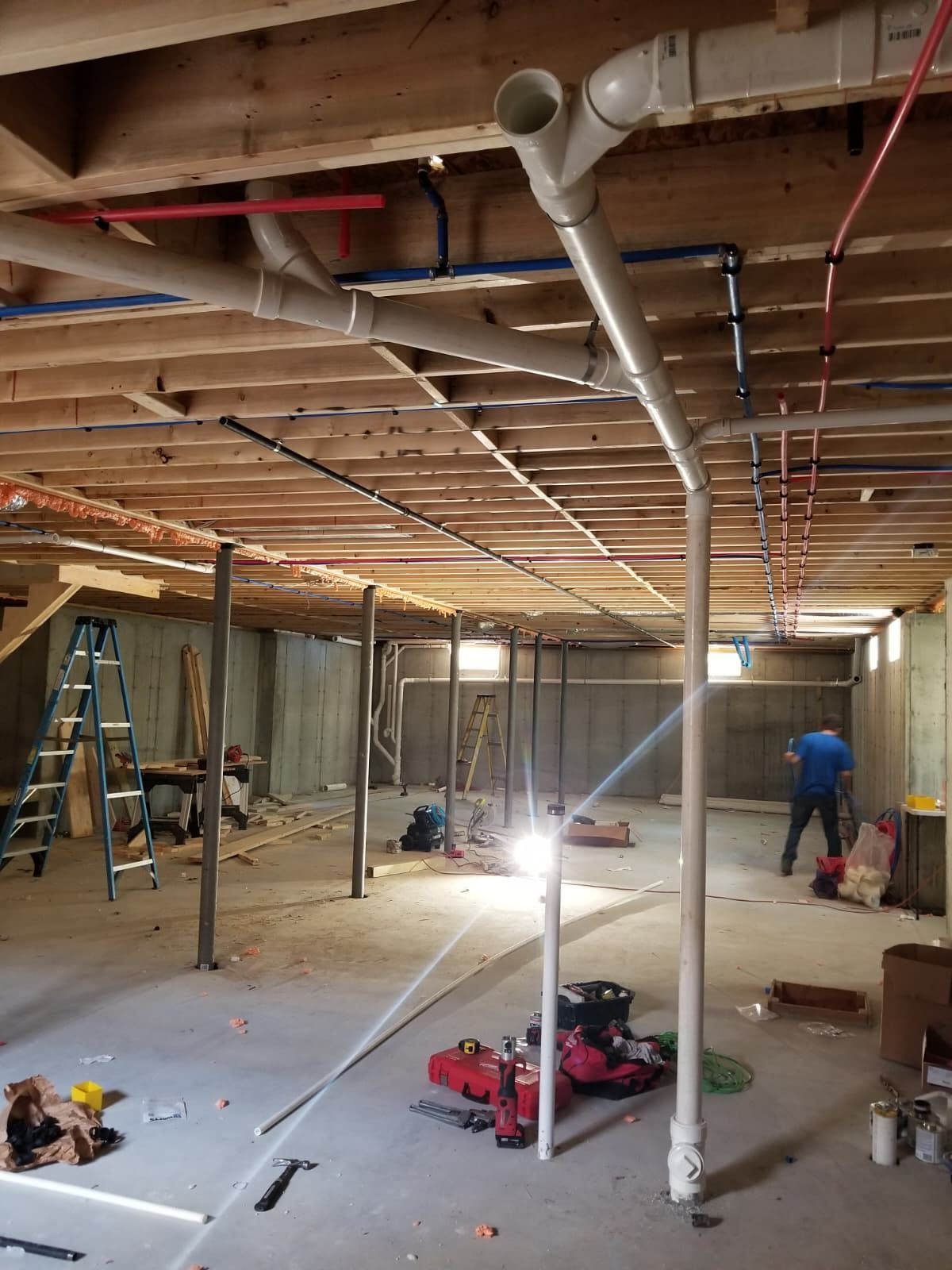 Basement under renovation with exposed beams, pipes, ladders, and two workers near bright lights
