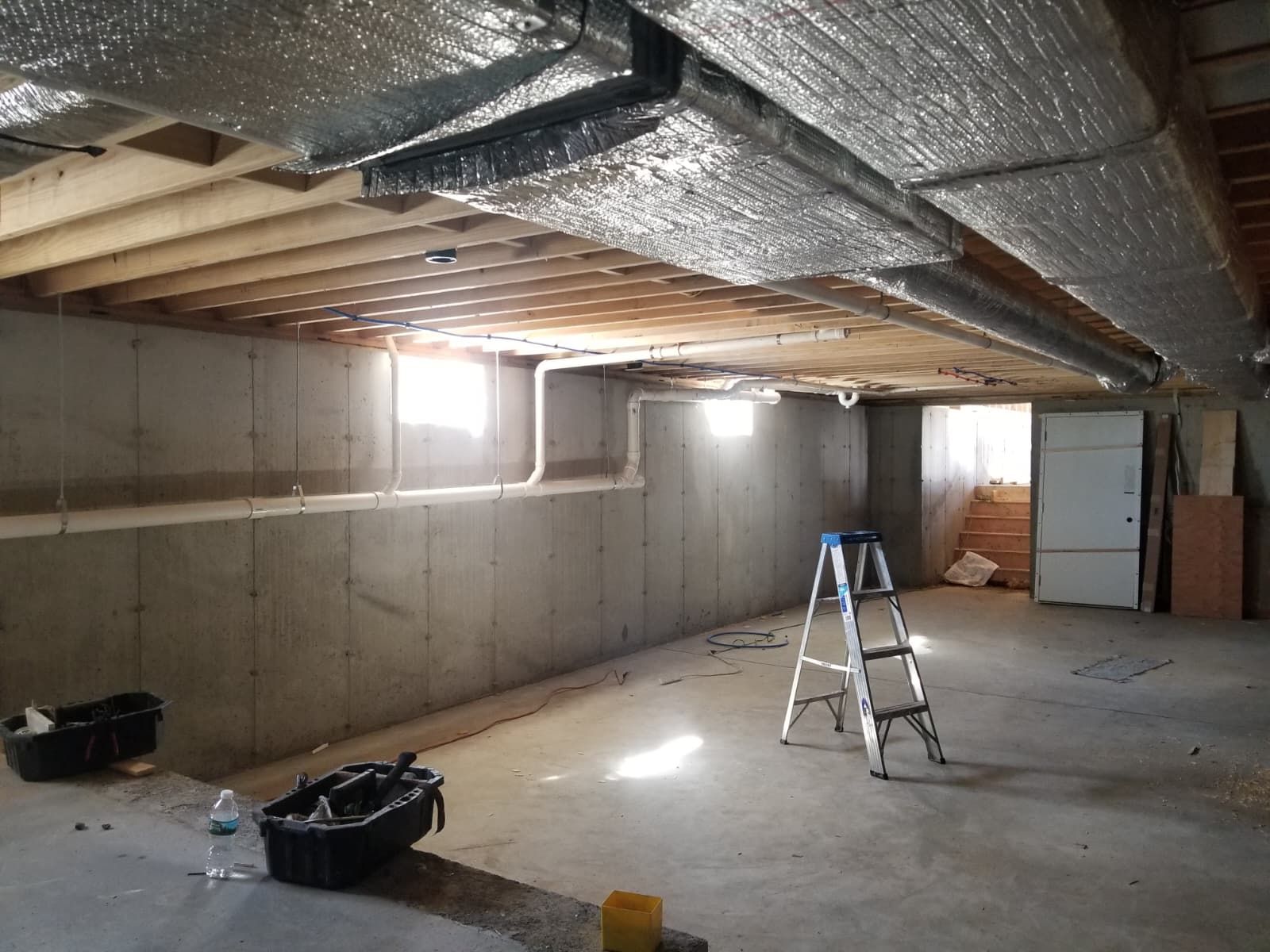 Unfinished basement with exposed insulation, concrete walls, ladder, storage bins, and a bright window.