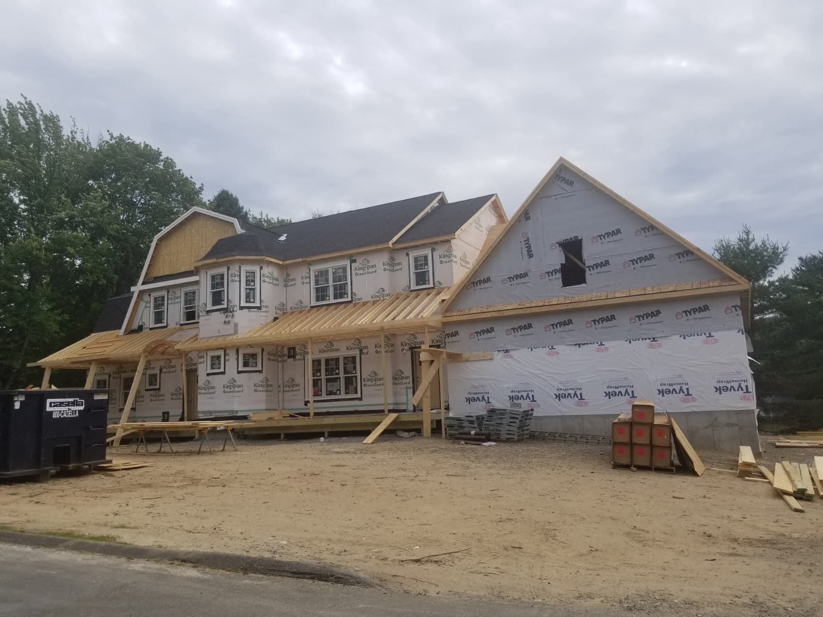 House under construction with wraparound porch, exposed framing, and white siding in a dirt lot.