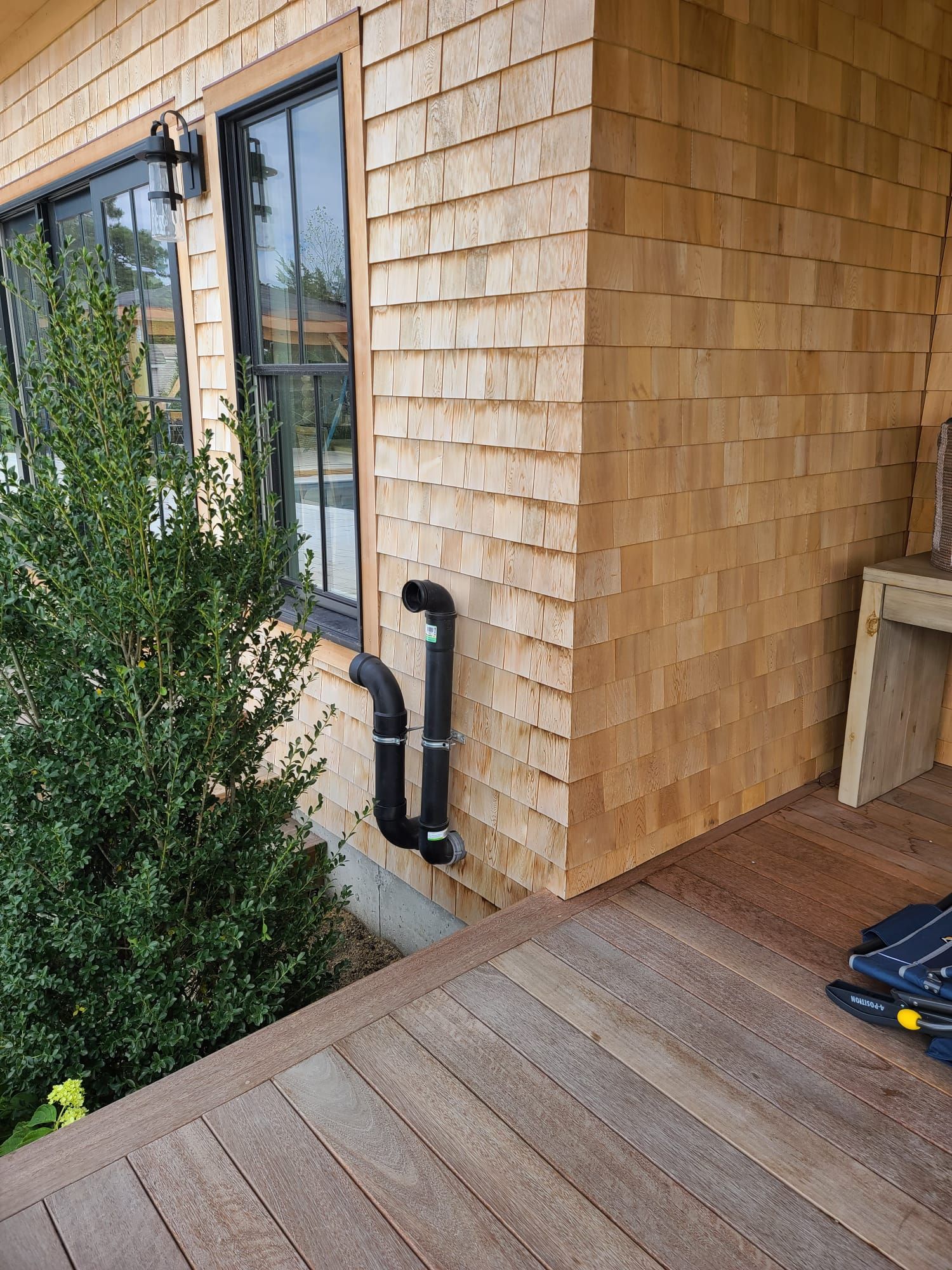 Wood-shingled house exterior with a door, black downspout, and wooden deck beside shrubs.