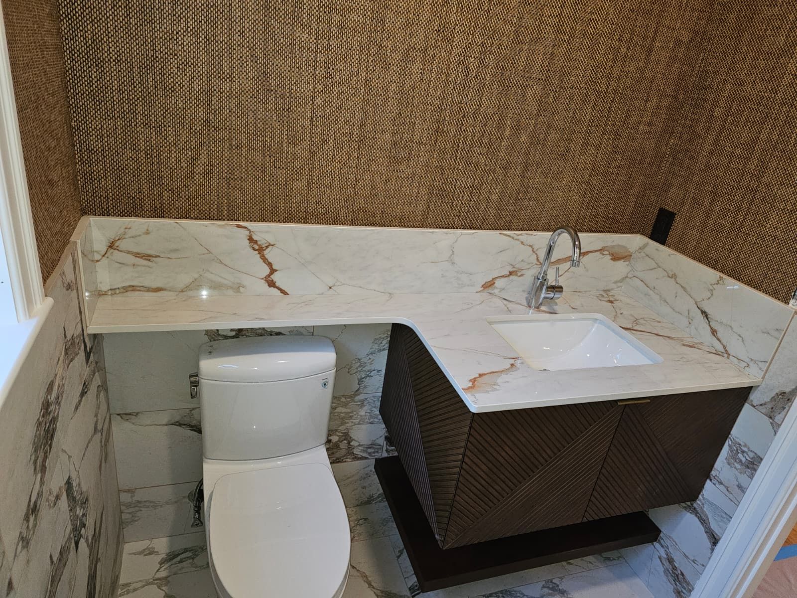 Compact bathroom vanity with white marble top, sink, and toilet beside woven wall panels