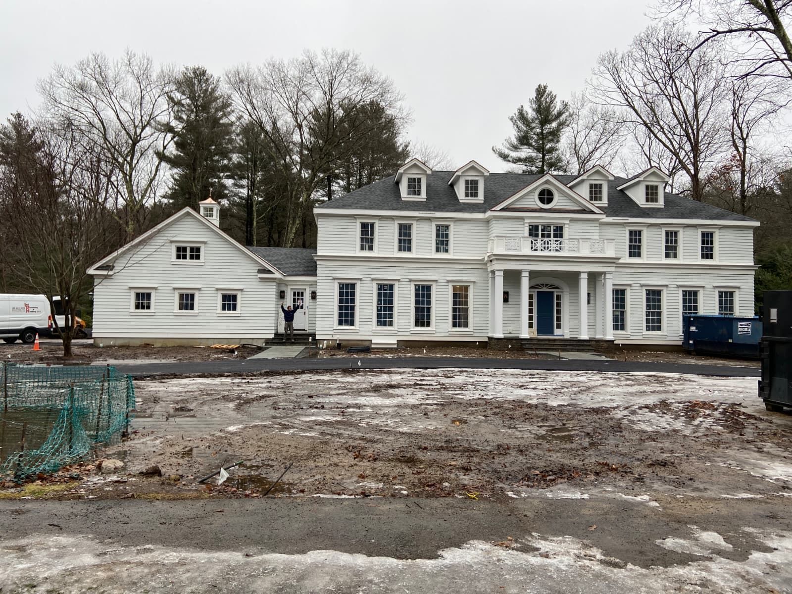 Large white house and smaller outbuilding on a muddy construction site under overcast skies