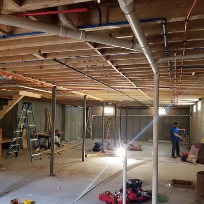 Basement under construction with exposed beams, ladders, tools, and two workers installing utilities