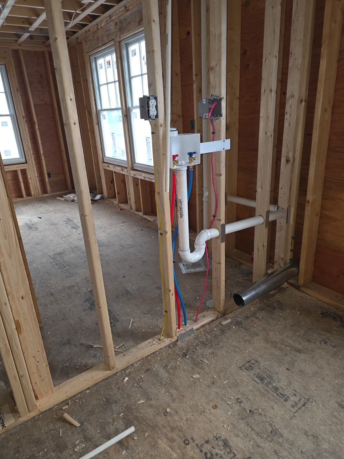 Unfinished house framing with exposed plumbing pipes and electrical wiring beside windows.