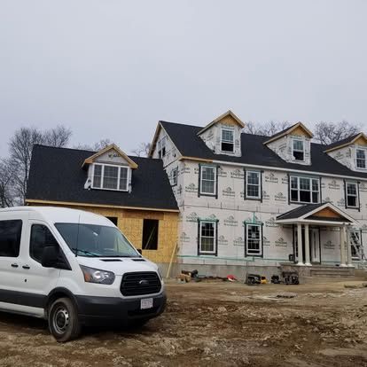 Construction site with white van and partially built house beside a larger unfinished townhouse under gray sky