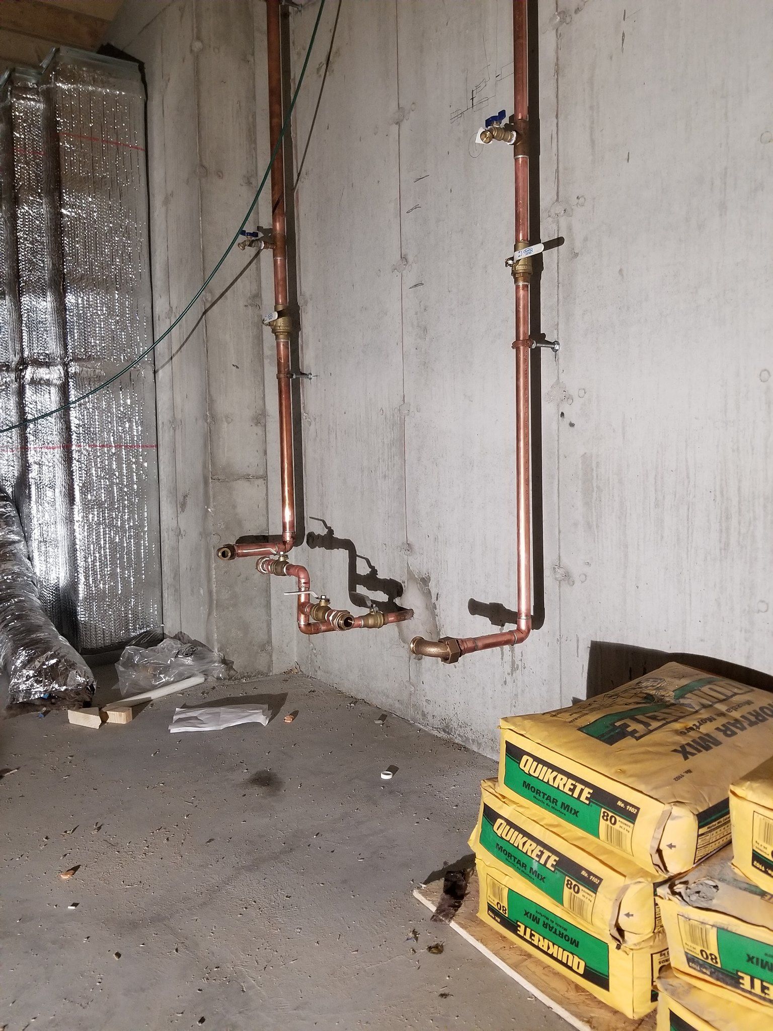 Basement wall with exposed copper pipes, insulation, debris, and stacked boxes of tile or flooring materials.