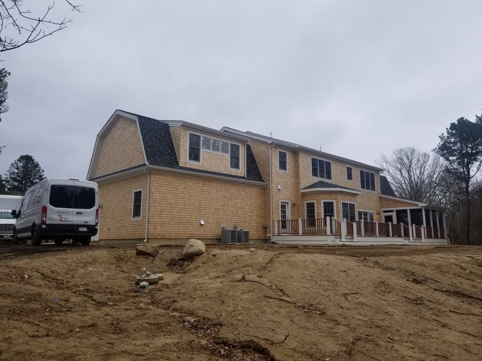 Large tan house with a white van parked beside it on a muddy, sloped lot under an overcast sky