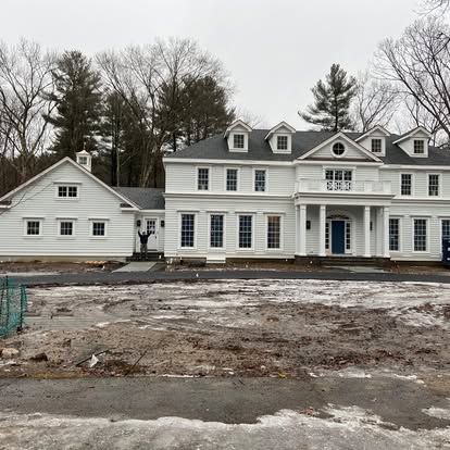 Large white house with dormer windows beside a smaller white building on a muddy, bare lot