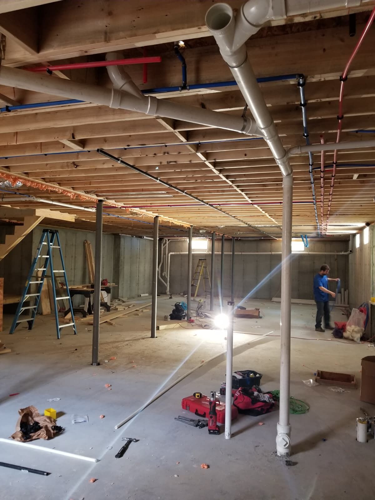 Unfinished basement with exposed joists, white pipes, tools, and two workers installing wiring and fixtures.