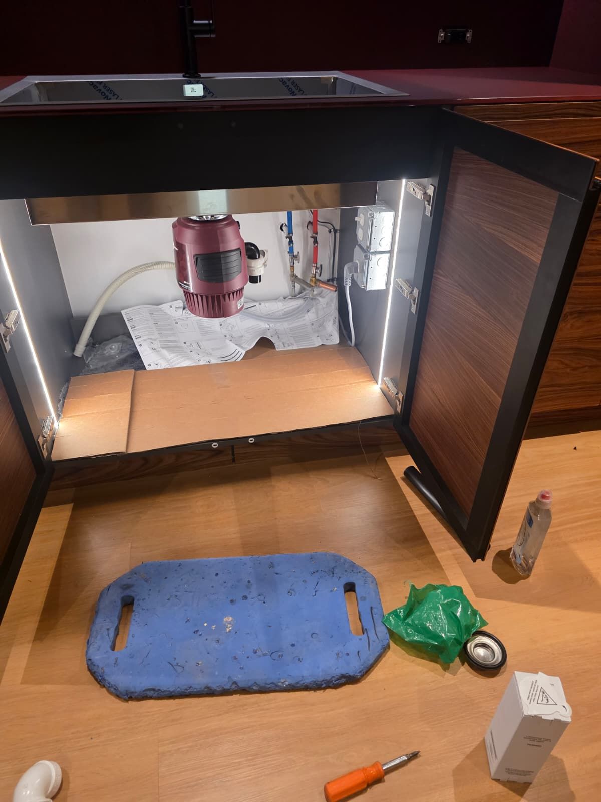 Open cabinet under a sink with tools and a blue mat on the floor, showing plumbing inside.