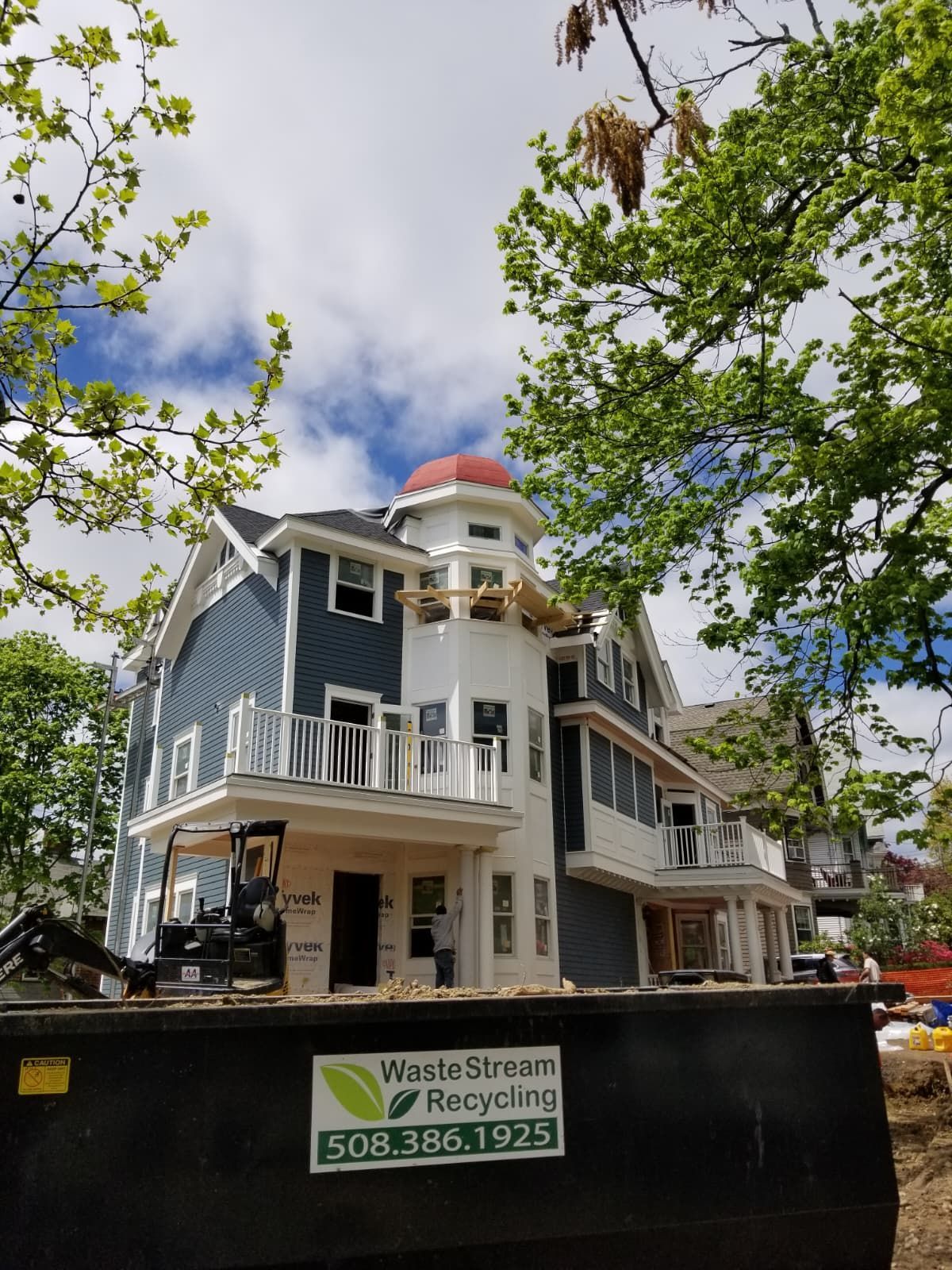 Partially built house with white trim behind a black WasteStream Recycling dumpster and trees