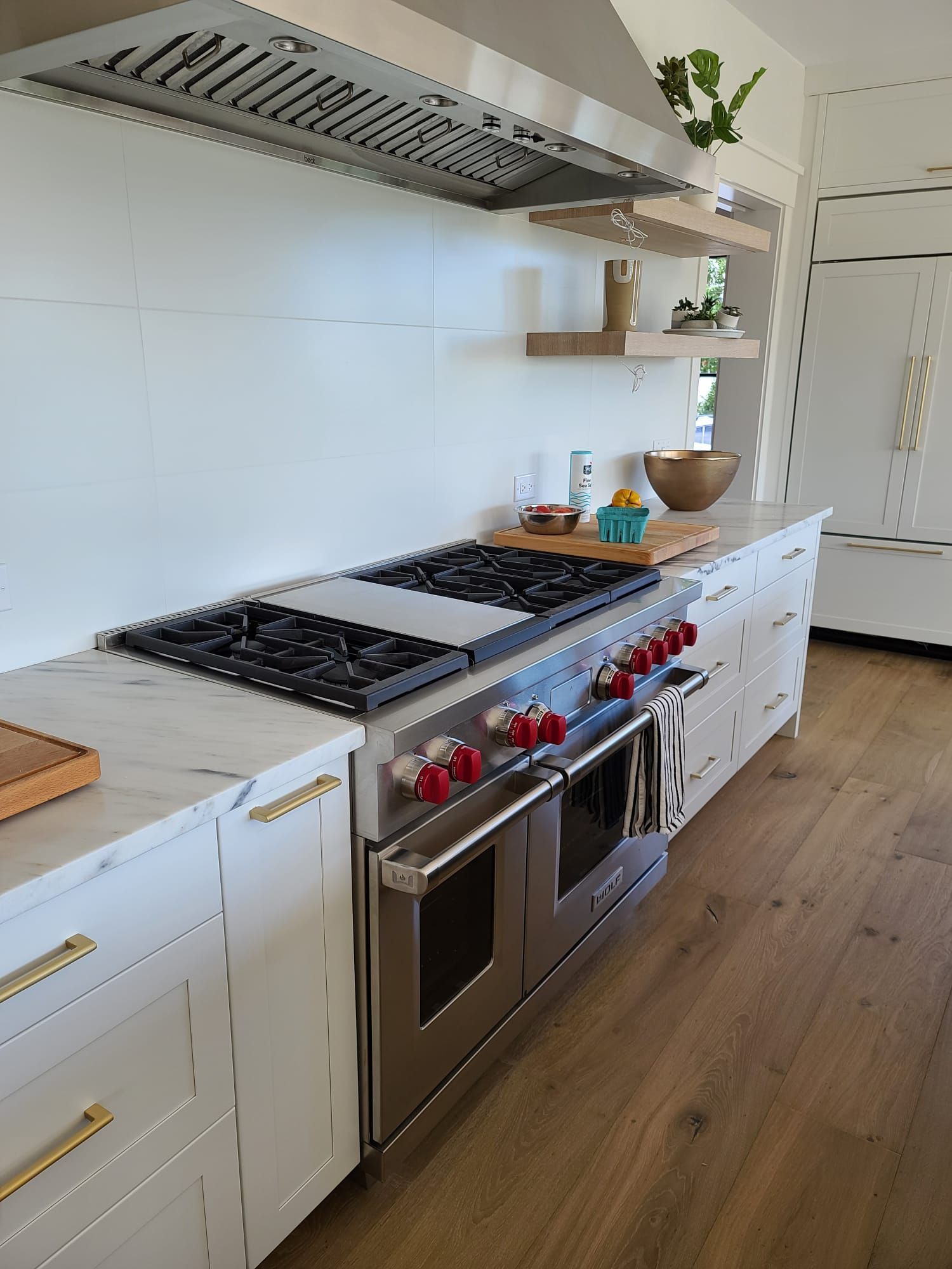 Modern white kitchen with stainless steel range, red knobs, open shelves, and wooden floors.