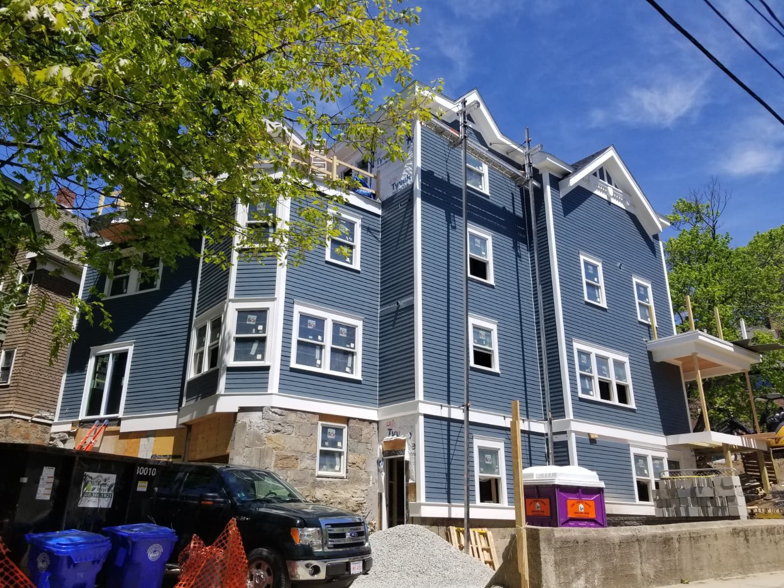 Blue multi-story residential building with white trim, trees, and parked cars on a sunny street