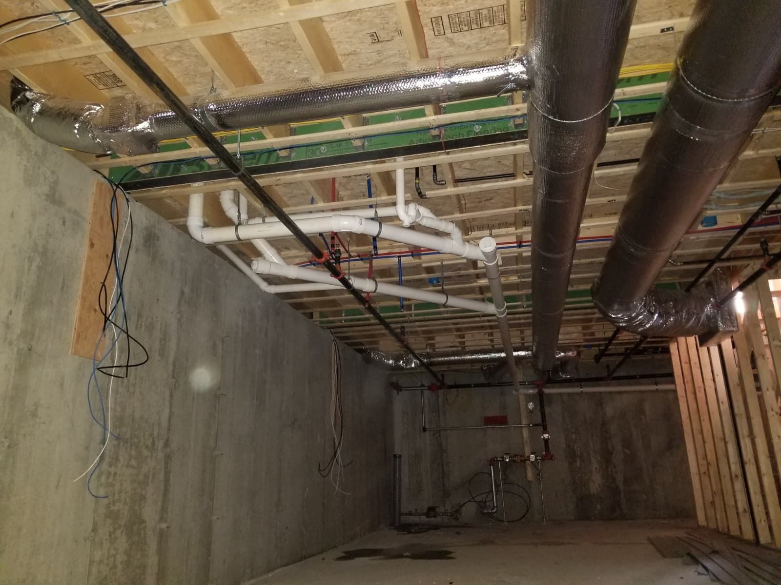 Unfinished basement ceiling with exposed pipes, wiring, and concrete walls under construction.