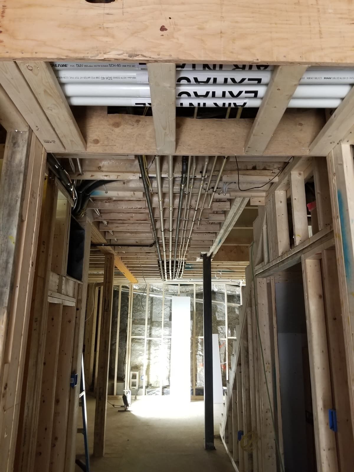 Unfinished hallway framing with exposed ceiling pipes and wiring in a construction site.