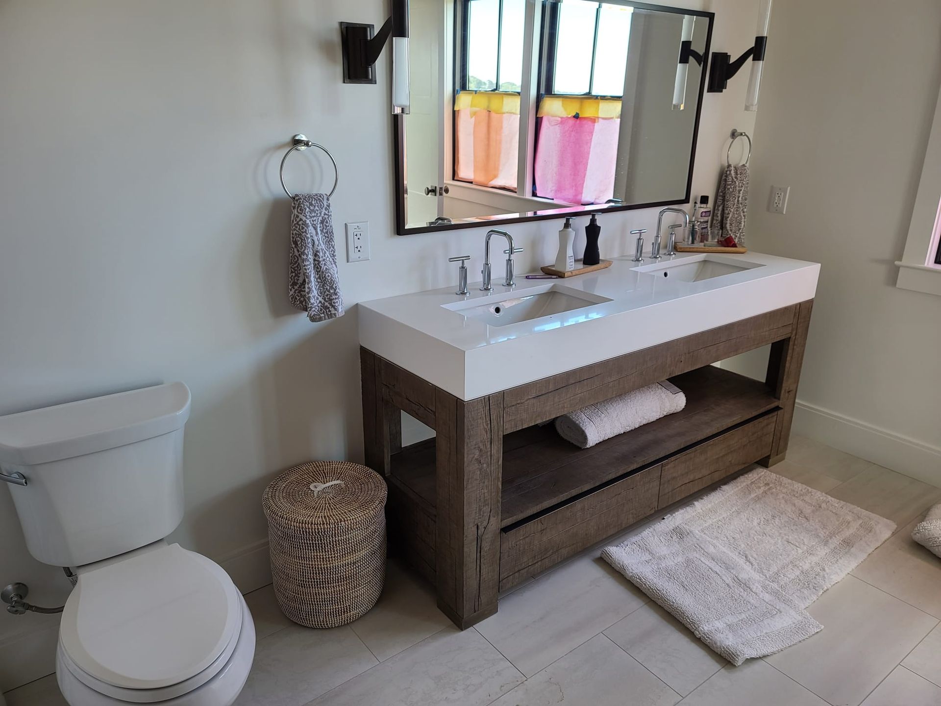 Bathroom with white toilet, double sink vanity, mirror, towels, and woven baskets on a light rug.