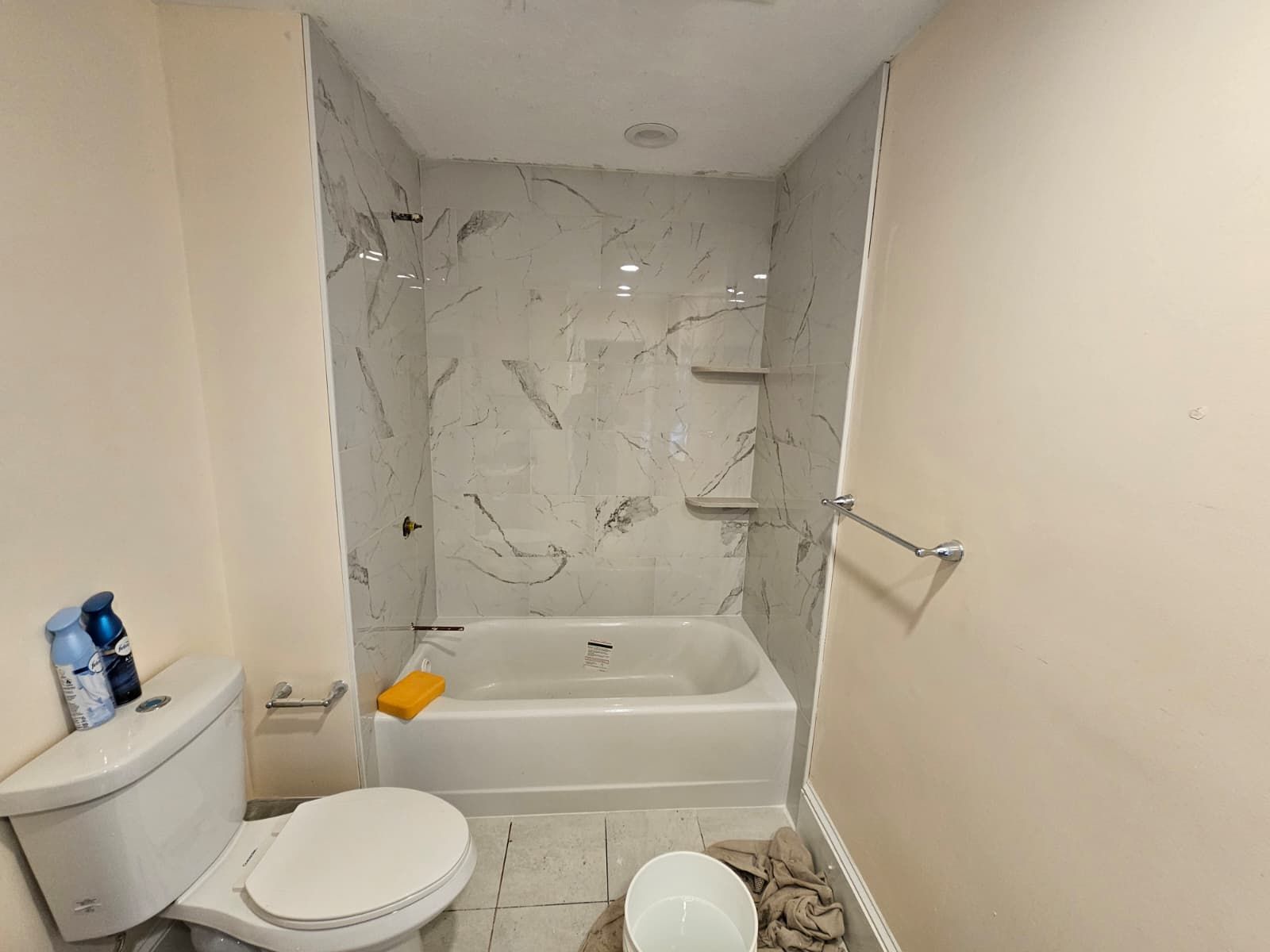 Small bathroom with toilet, bathtub, and marble-pattern shower wall; unfinished floor tiles and a grab bar.
