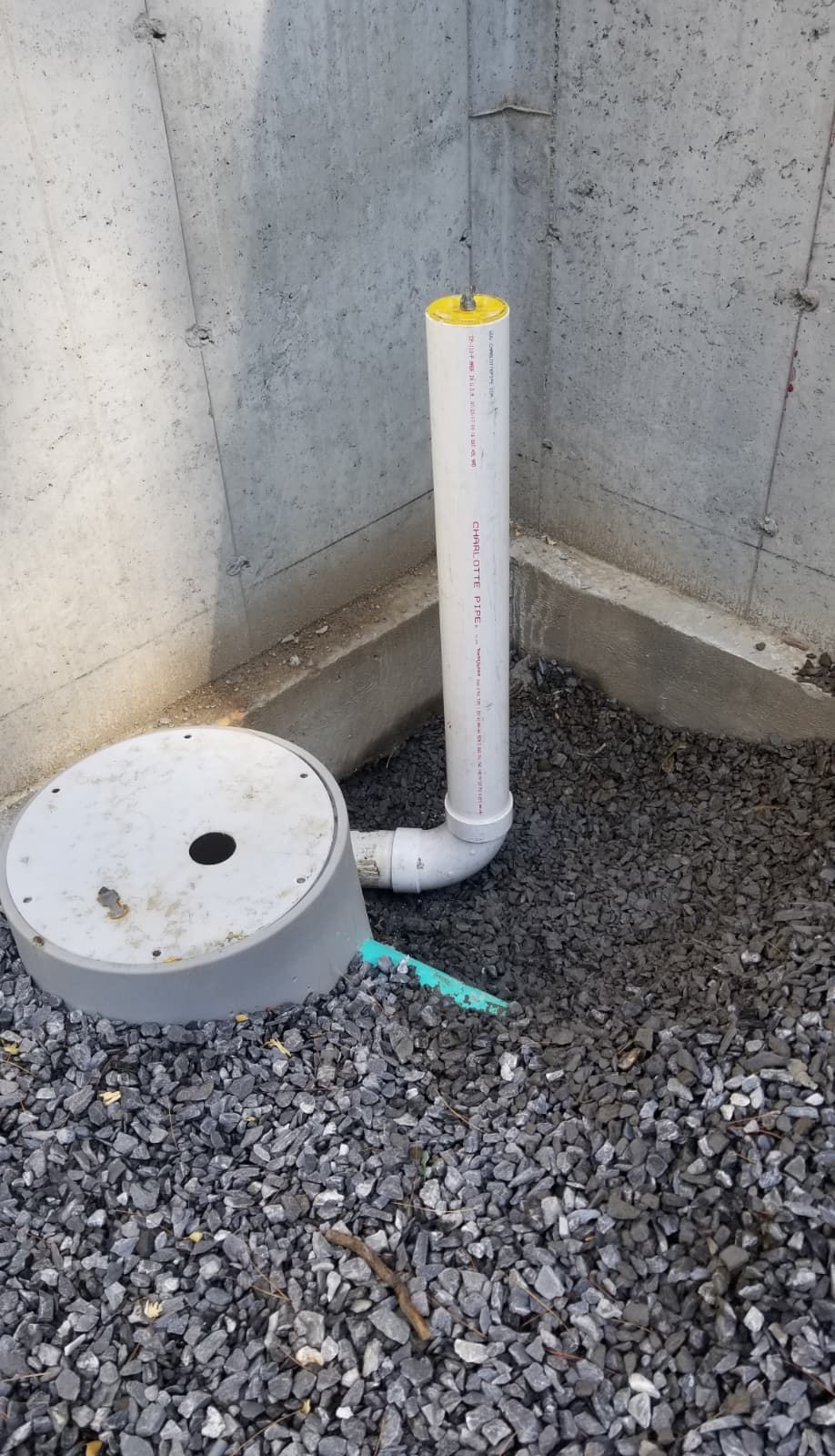 White PVC pipe and capped drain cover in gravel beside a concrete wall corner.