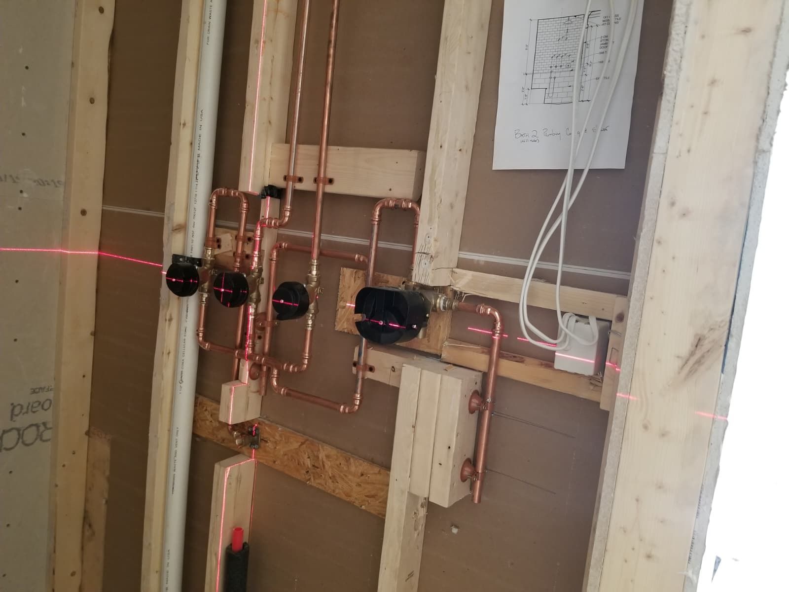 Plumbing valves and copper pipes inside an unfinished wall, with exposed wiring and a paper note nearby