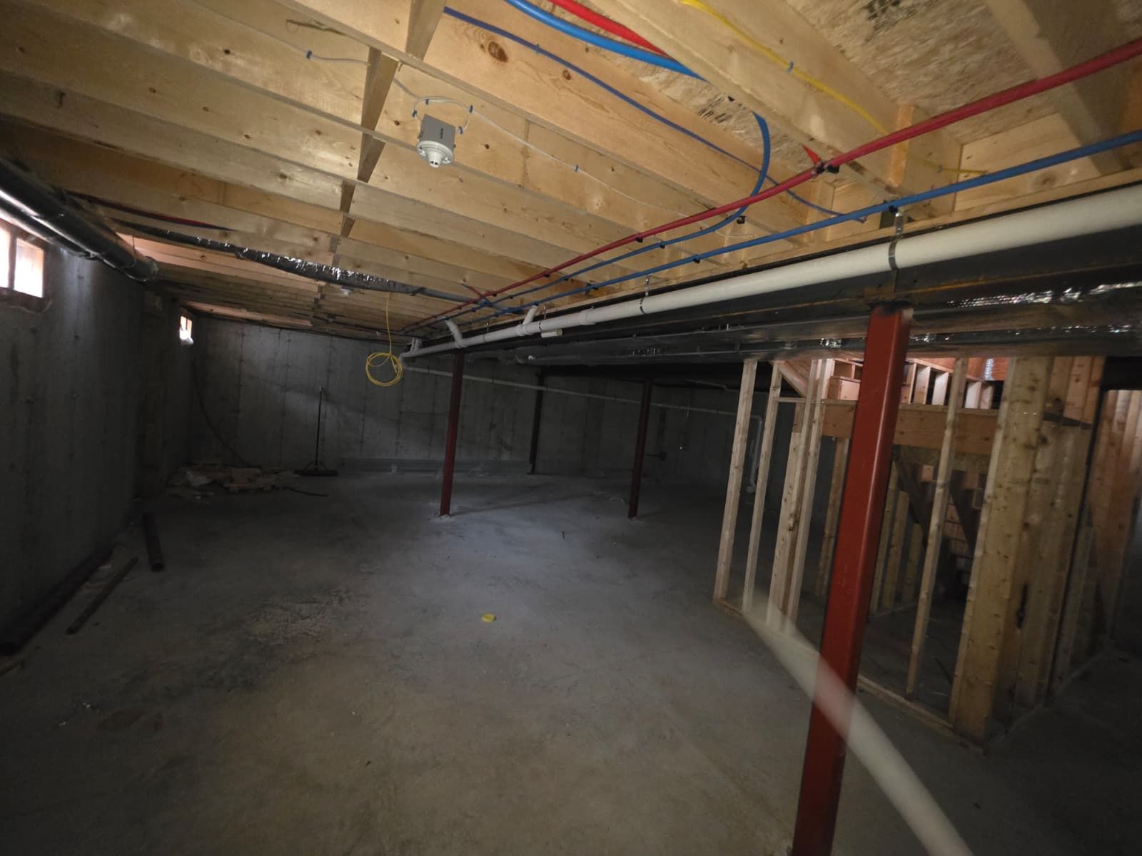 Unfinished basement with exposed wooden beams, concrete floor, and visible plumbing and wiring.