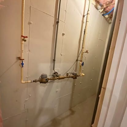 Utility pipes and valves mounted on an unfinished basement wall.