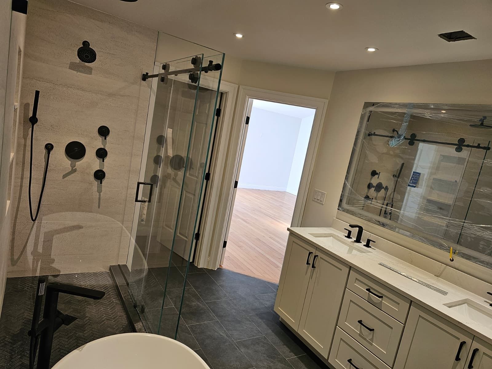 Modern bathroom with glass shower, vanity, large mirror, and dark tile floor, opening to a bright room