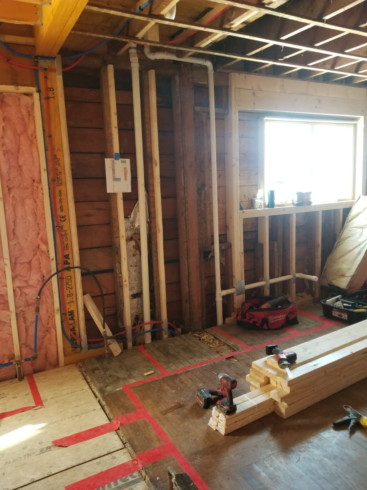Interior under renovation with exposed studs, red tape on floor, window light, and tools on the floor