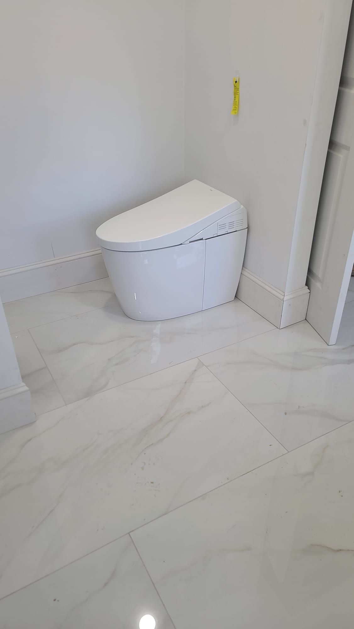 White toilet in a tiled bathroom corner with a yellow cleaning tag on the wall