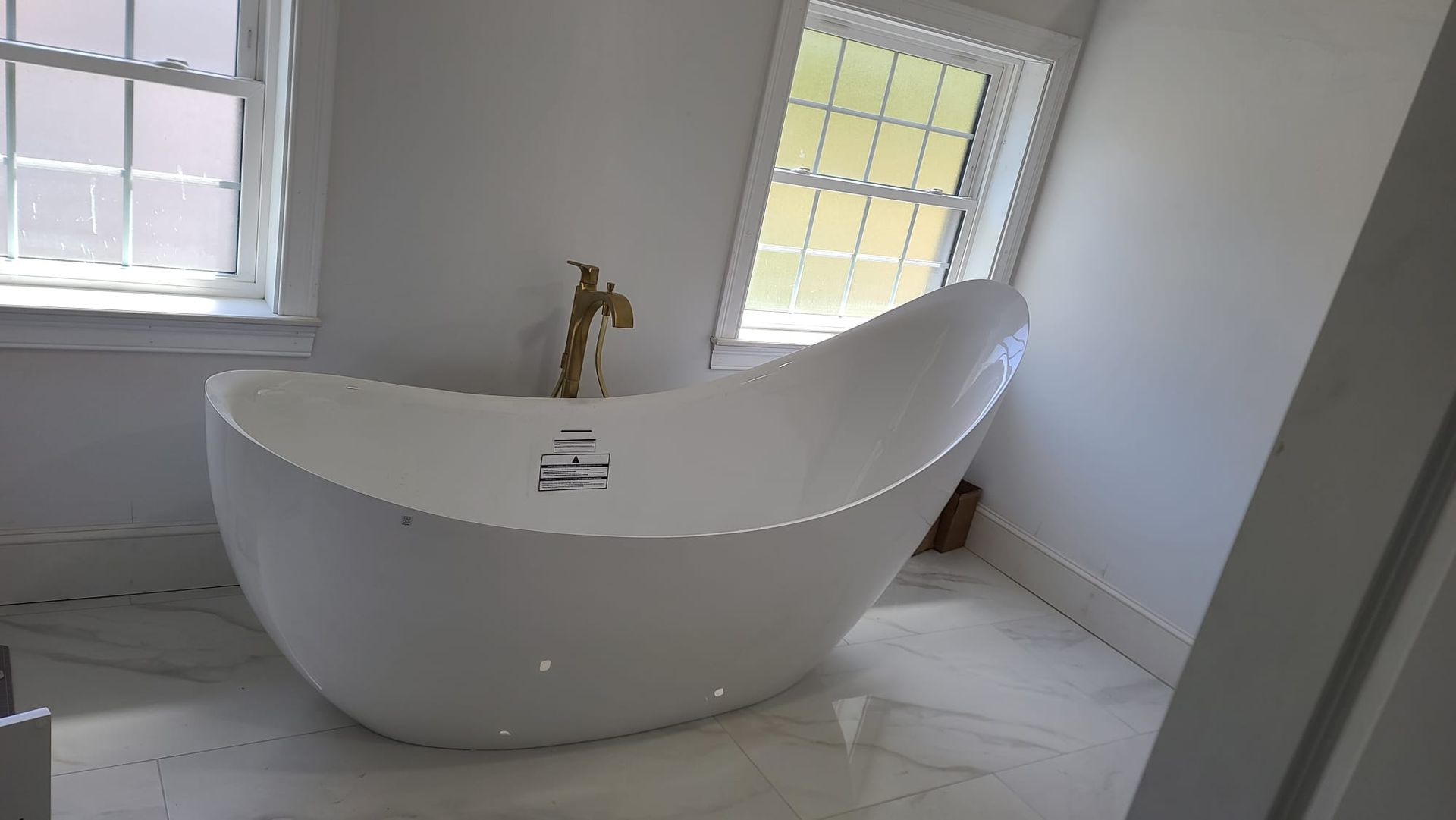 Modern freestanding white bathtub in a bright bathroom with windows and gold faucet