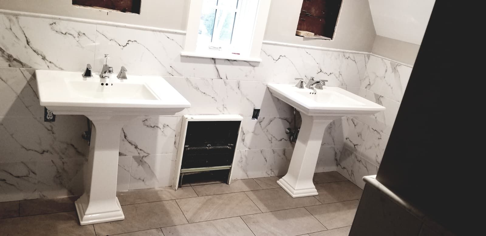 Two white pedestal sinks in a tiled restroom with marble-pattern walls and a window above.