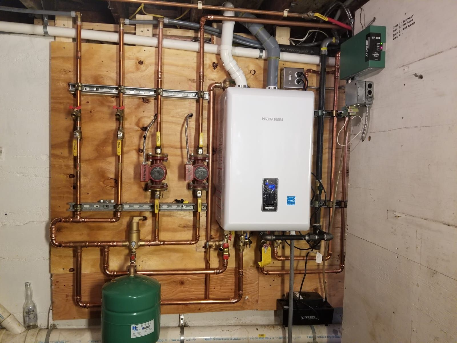 Boiler room with wall-mounted white heater, copper pipes, green expansion tank, and controls on a concrete wall