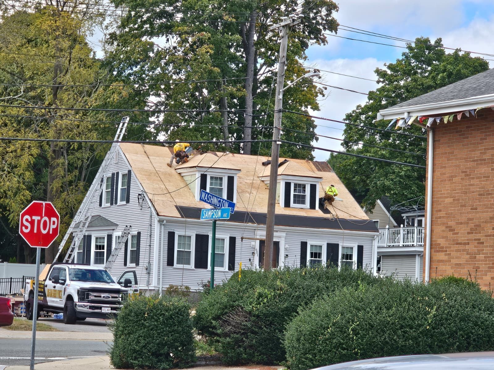 Residential street with a white house under roof repairs, stop sign, parked cars, and trees.