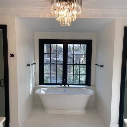 Bright white bathroom with a freestanding tub beneath a black-framed window and chandelier above.
