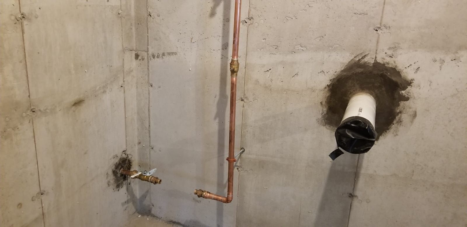 Concrete basement wall with exposed copper pipes and a circular drain/vent opening