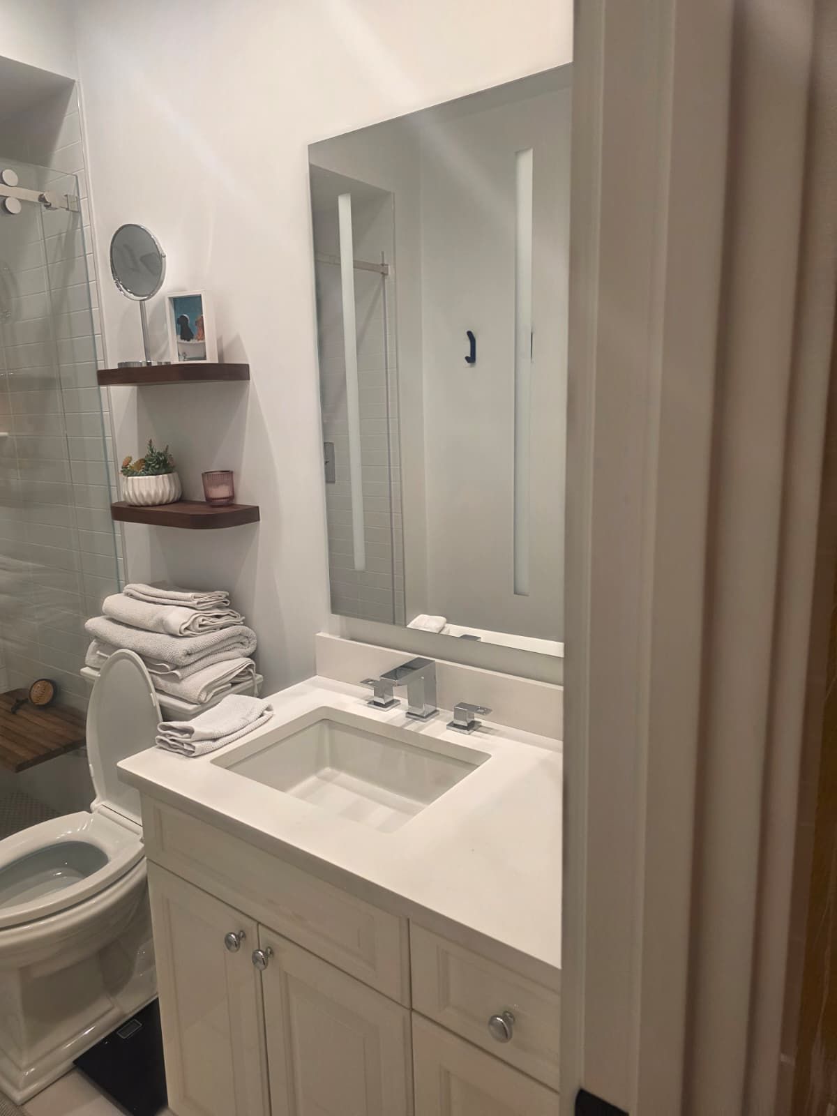 Compact bathroom with white vanity, mirror, sink, toilet, and shelves of folded towels.