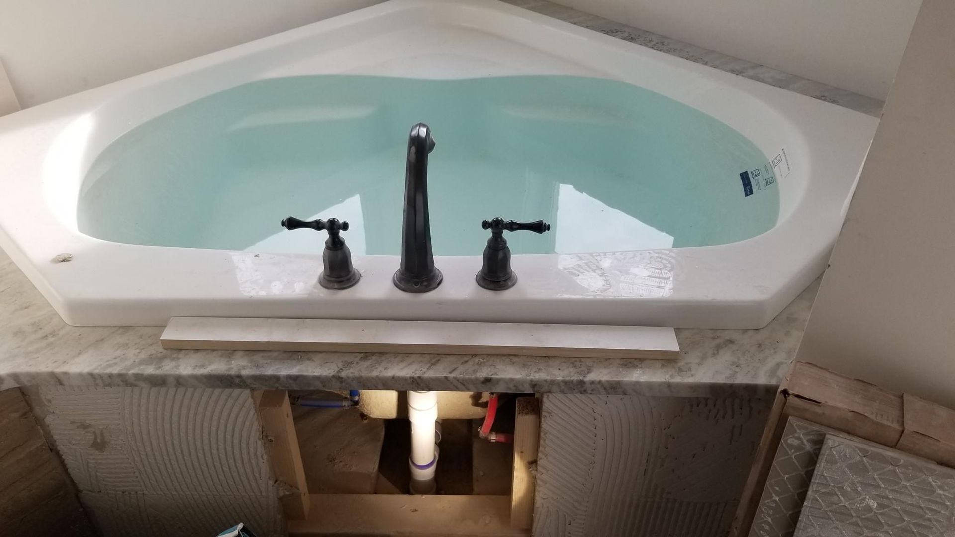 Built-in bathtub with pale blue water and black faucet fixtures in an unfinished bathroom wall opening