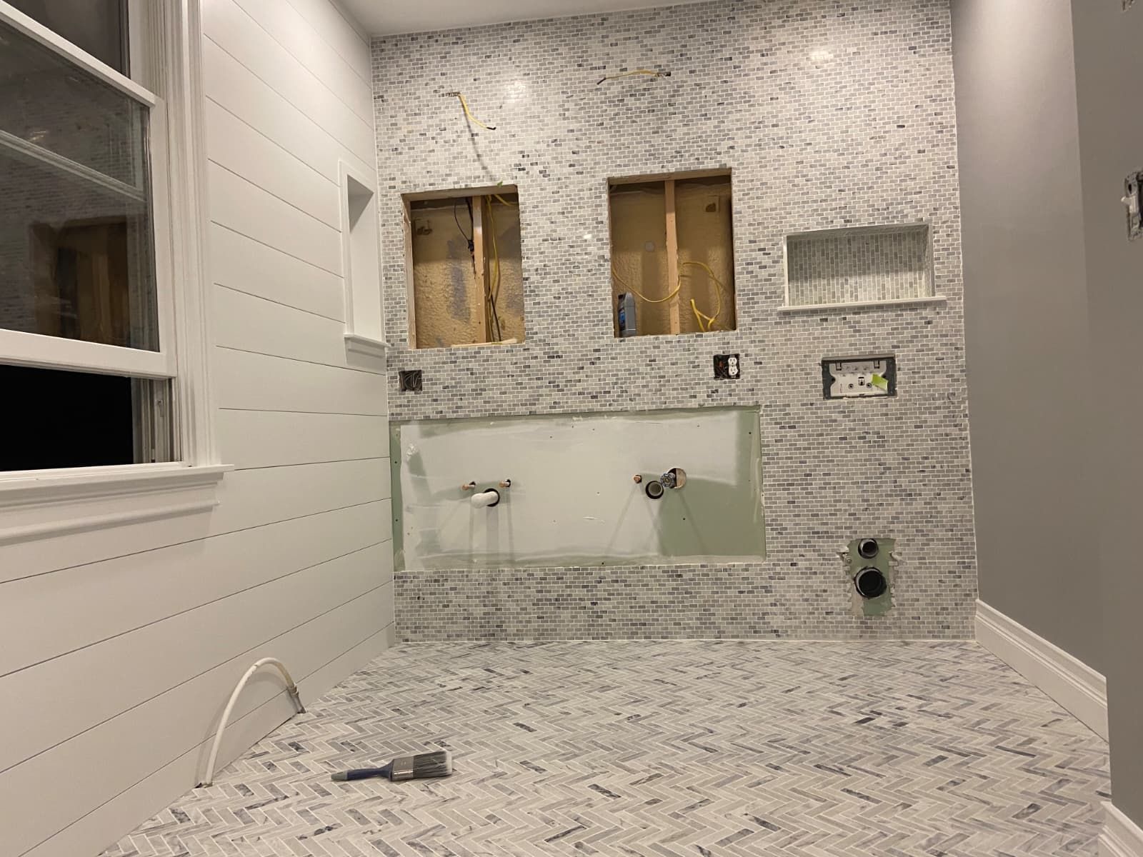 White-tiled bathroom under renovation with a sink, mirror, and exposed wall openings above it