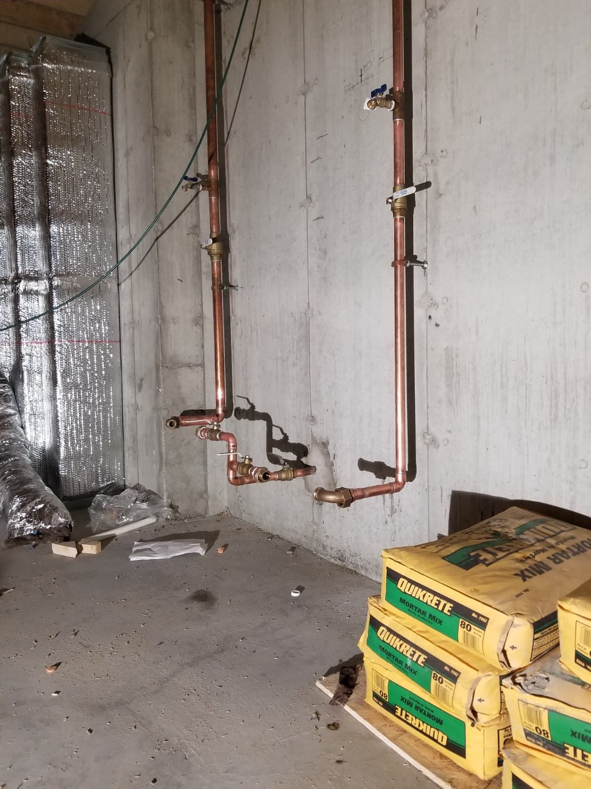 Unfinished basement wall with exposed copper pipes, insulation, and stacked cardboard boxes on the floor