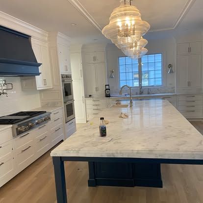 Bright white kitchen with a large marble island, blue range hood, and hanging glass chandeliers.