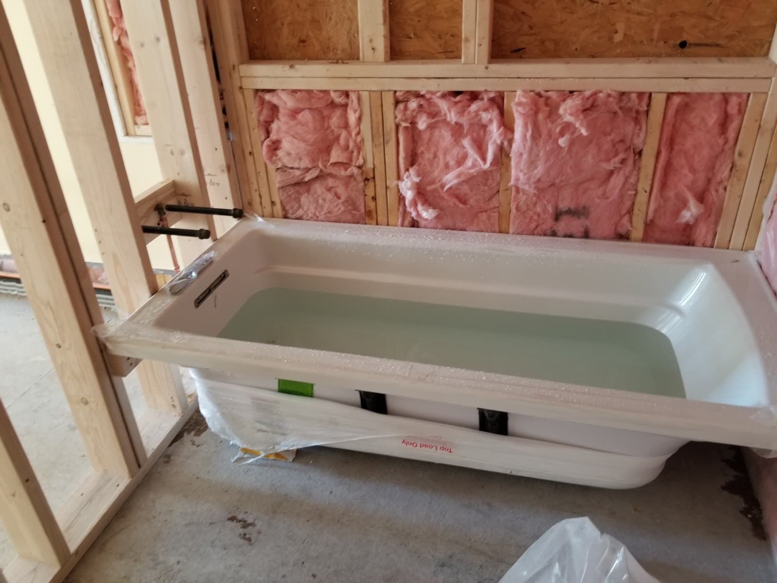 Freestanding white bathtub installed in an unfinished bathroom with exposed pink insulation and wood framing