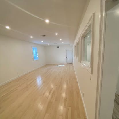 Empty modern room with light wood floors, white walls, recessed lights, and windows along one side