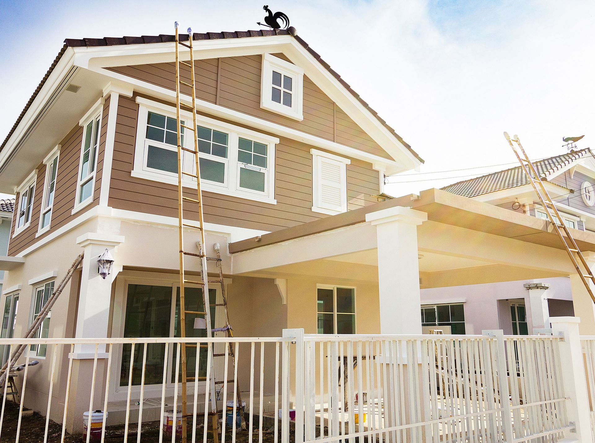 A two-story house with tan shingles and cream trim, featuring a front carport and two ladders leaning against the exterior.