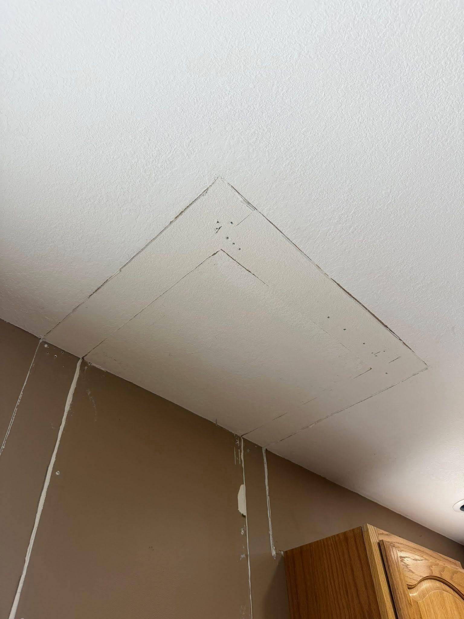 A ceiling with a marked square, possibly for repair. Brown wall and part of a cabinet visible below.